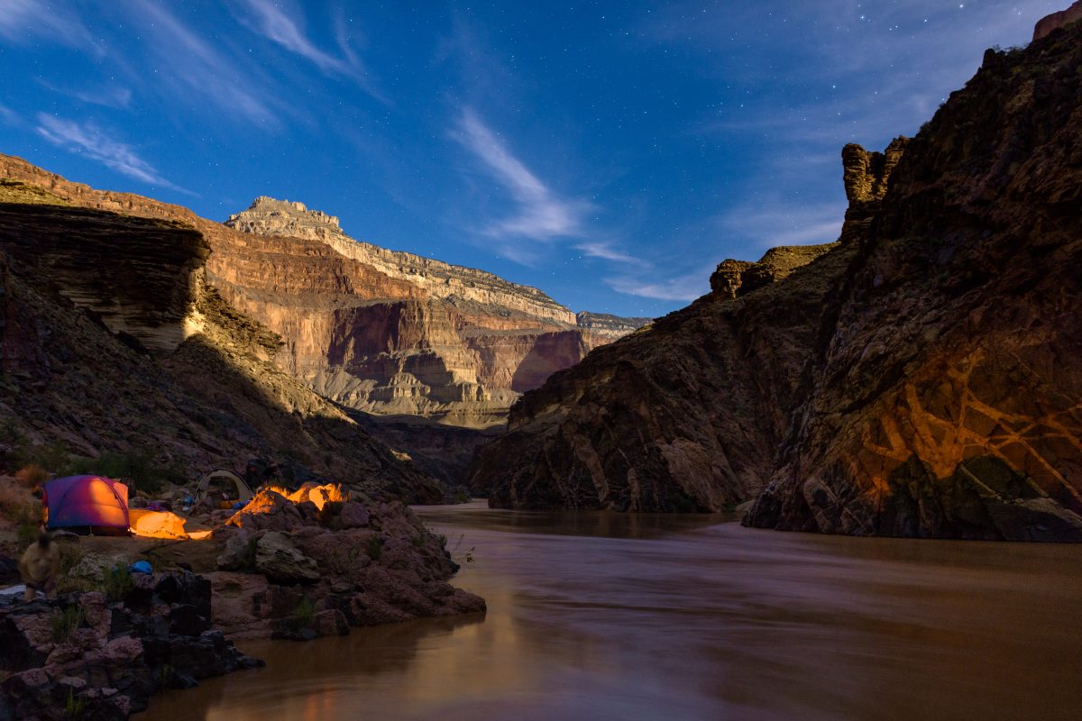 A nighttime view of the Grand Canyon from the Colorado River, with stars above and a campsite glowing on the riverbank.